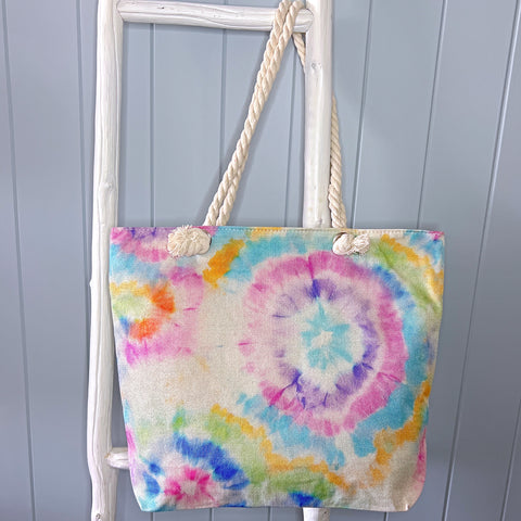 Personalised beach bag or beach tote hanging from a white timber ladder with its long rope handle. The bag has a tie dye pattern in pastel shades of the rainbow.