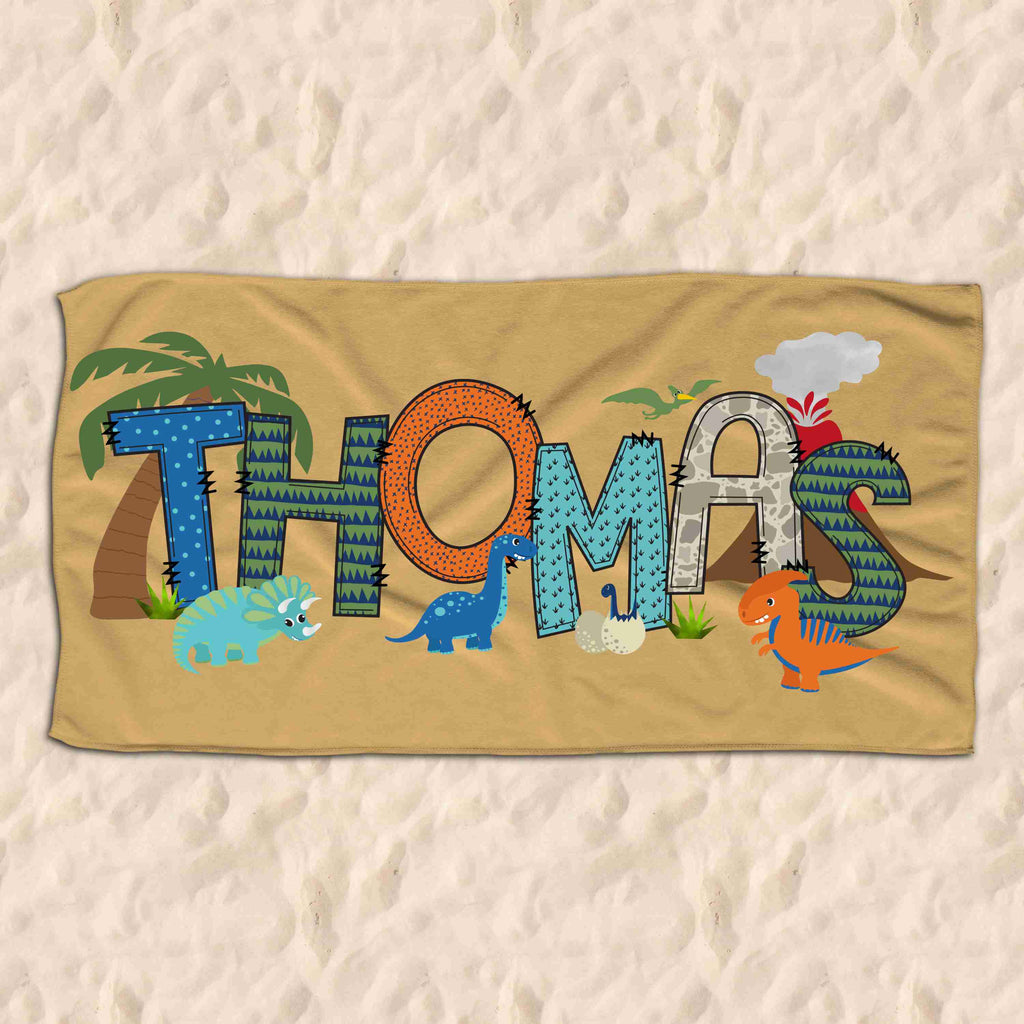 Beach towel on sand. Beach towel is personalised with the name Thomas in dinosaur themed letters. It also includes various graphics of dinosaurs, trees and volcanoes.