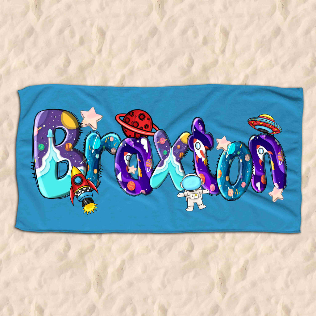 Personalised childrens beach towel lying on a sandy beach. The beach towel is blue personalised with the name Braxton in a space themed doodle font.