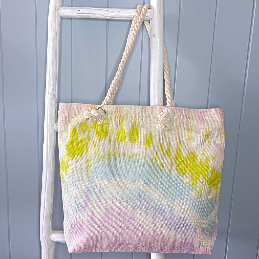 Personalised beach bag or beach tote hanging from a white ladder using its rope handle. The bag has a tie dye pattern in shades of pink, green and blue.