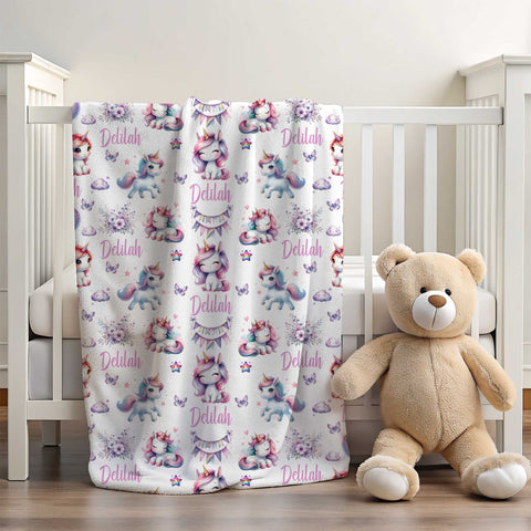 White wooden cot with white minky blanket draped over side next to a beige coloured teddy bear. The personalised minky fleece blanket has various rainbow coloured unicorns in different poses amongst clouds, flowers, stars and butterflies.