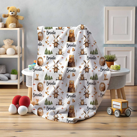 A personalised white minky fleece blanket is draped over a small table within a nursery setting. The personalised minky fleece blanket has various woodland animals among trees, leaves and mushrooms, personalised with the name Bronte.