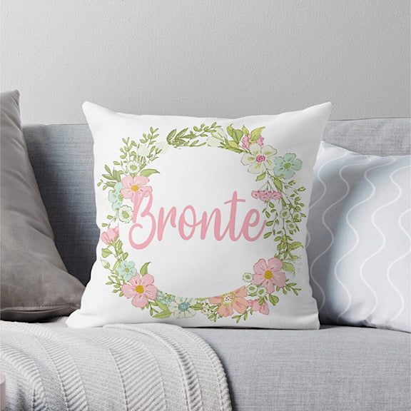 White cushion with a ring of pastel coloured flowers personalised with a name in light pink sitting on a grey couch