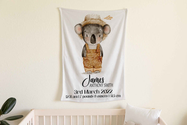 A white fleece minky blanket personalised with a name and birth details below a watercolour picture of a grey koala wearing a straw hat and brown coloured overalls like a farmer. The blanket is hanging in a nursery above a timber cot.