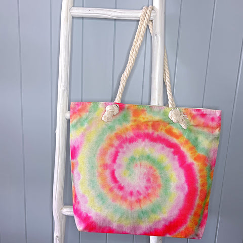 Personalised beach tote or beach bag hanging from a white timber ladder using its rope handles. The bag has a tie dye pattern in bright shades of the rainbow.