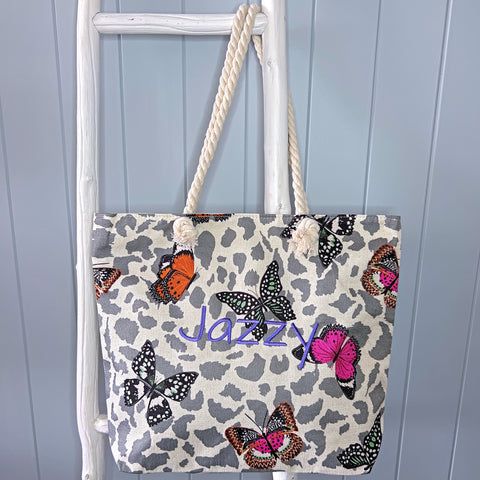 Beach bag or tote bag hanging from a white ladder. Bag has grey leopard spots in the background with different coloured butterflies on top, with the name Jazzy embroidered in dark purple on the front. Bag has rope handles.