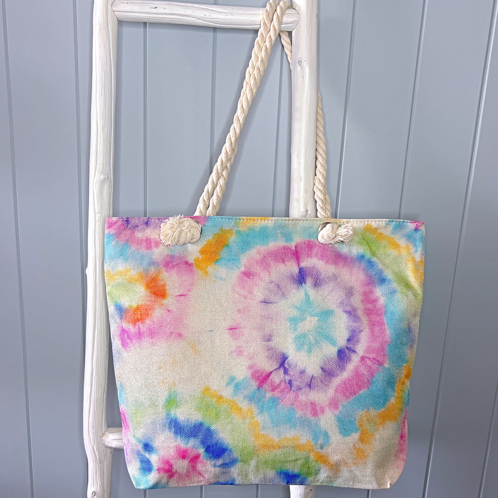 Personalised beach bag or beach tote hanging from a white timber ladder with its long rope handle. The bag has a tie dye pattern in pastel shades of the rainbow.