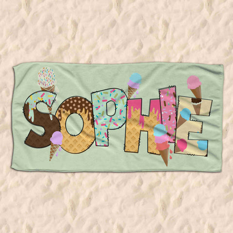 Mint green childrens beach towel lying on the sound. The towel has the name Sophie on it in ice creamed themed letters.