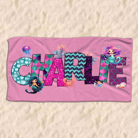 Personalsied pink childrens beach towel resting on sand. Beach towel has the name Charlie written on it in a mermaid themed font.
