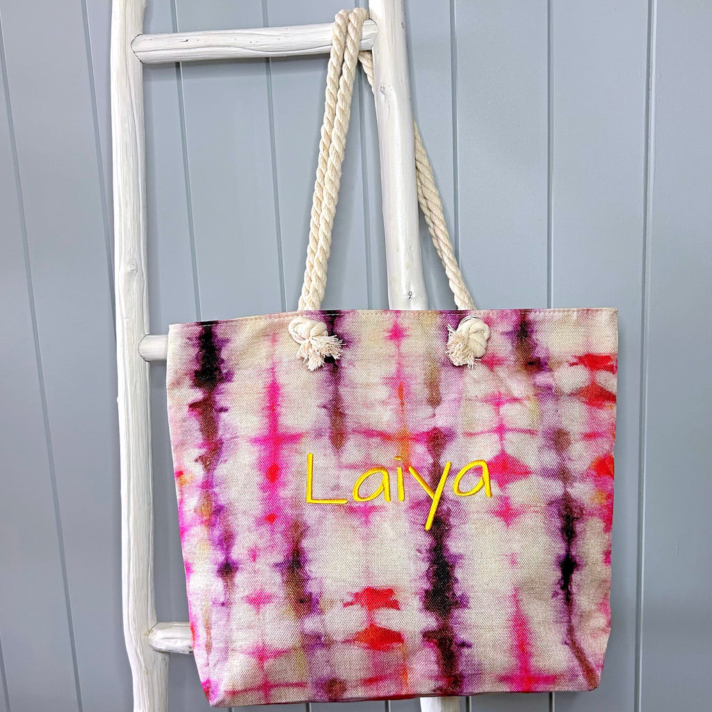 Beach bag or tote bag hanging from a white ladder. Bag is a red, pink, purple and orange tie dye pattern with the name Laiya embroidered in yellow on the front. Bag has rope handles.