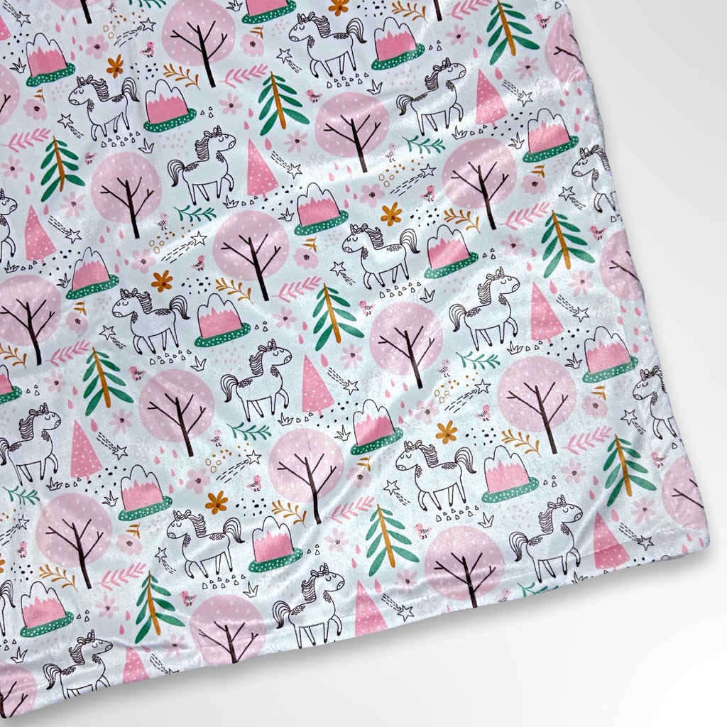 Minky blanket with white unicorns prancing through a pastel pink and green forest on a white background, ready to be personalised with a name in embroidery