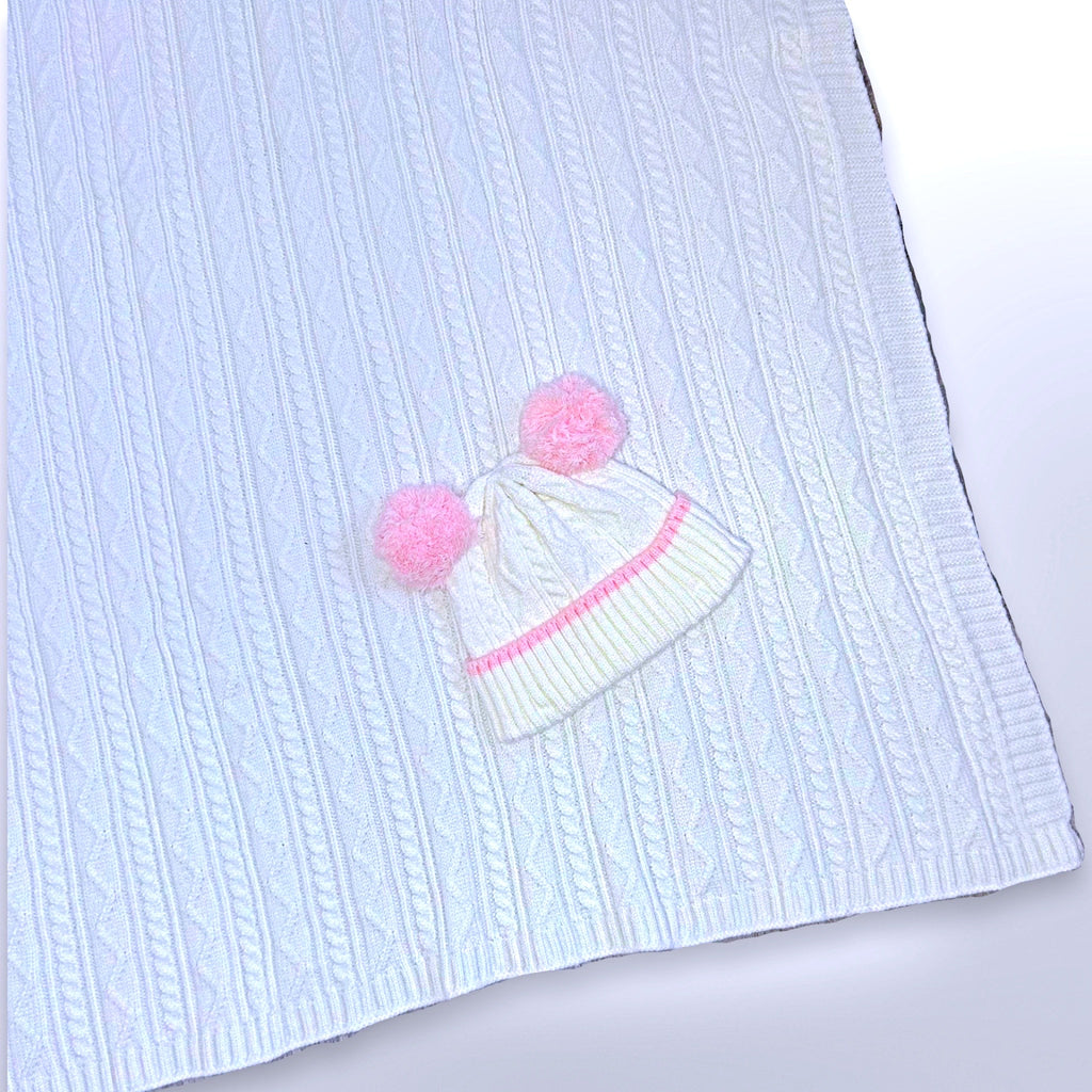 White cable knit blanket with matching beanie that has pink edging and pink poms poms, ready to be personalised with a name using embroidery.