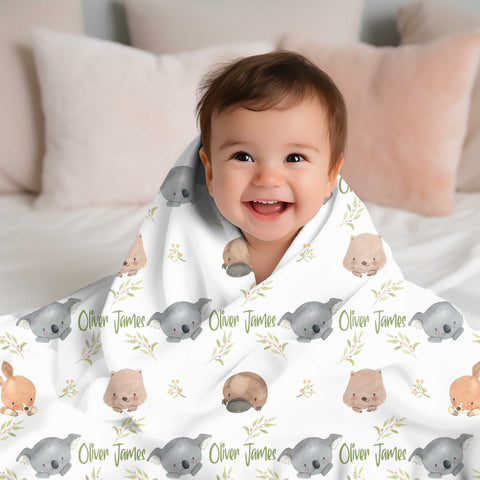 Smiling baby with brown hair wrapped in a personalised minky blanket. The blanket has australian animals and wattle on it along with the name Oliver James.
