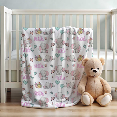 White wooden cot with personalised minky blanket draped over one side with tan teddy bear sitting next to it. Personalised minky blanket has grey elephants with large pink ears in various positions holding mint green heart shaped balloons. The background of the blanket also has pink and mint coloured hearts on it.