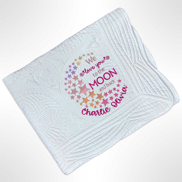 White heirloom quilt blanket with scalloped edges. In the bottom right of the blanket there is an embroidered design that has stars in various shades of pink and purple in an arc with the saying We Love you to the Moon and Back in the middle of the stars and with the name personalised underneath.