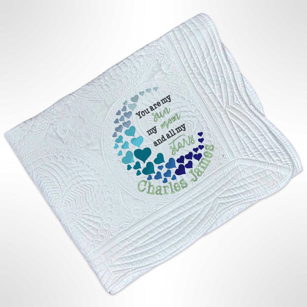 White heirloom quilt blanket with scalloped edges. In the bottom right of the blanket there is an embroidered design that has hearts in various shades of blue and green in an arc with the saying You are my sun my moon and all my stars in the middle of the hearts and with the name personalised underneath.