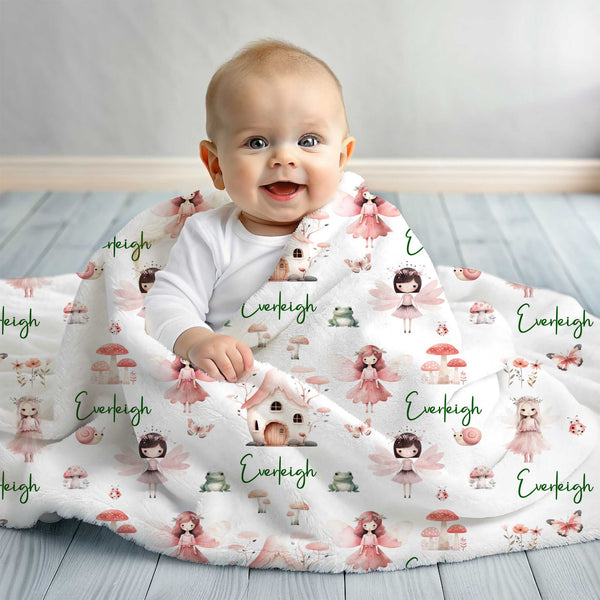 A baby sitting up wrapped in a personalised minky fleece blanket. The blanket has fairies in a fairy garden all in pink shades.