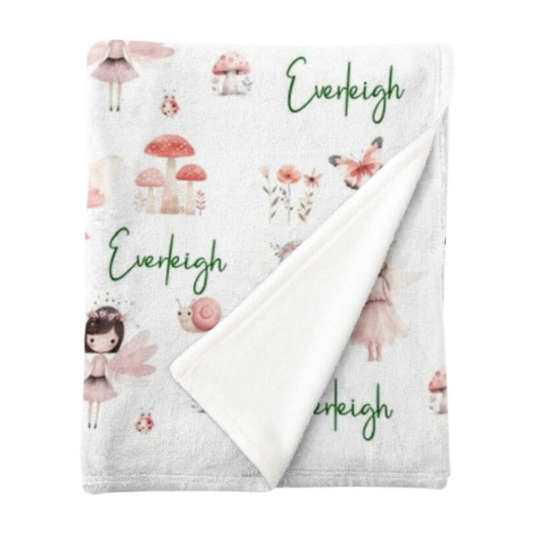 A folded personalised minky fleece blanket covered in fairies in a fairy garden in pink shades.
