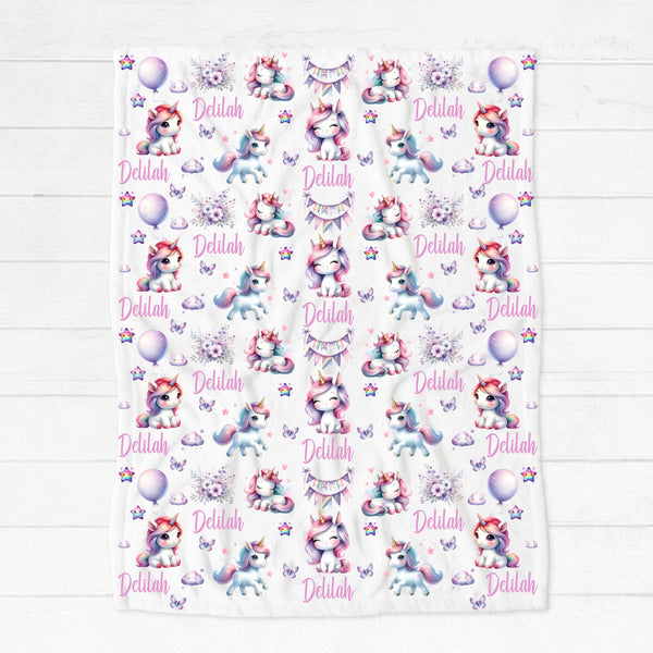 White minky fleece blanket placed on white floorboards. The blanket has white unicorns with rainbow coloured manes among pastel coloured flowers, clouds, butterflies and stars.