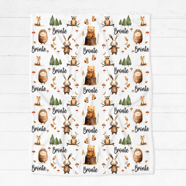 A personalised minky blanket lying on a white wooden floor. The blanket has various woodland creatures among trees, leaves and mushrooms in neutral shades, personalised with the name Bronte.