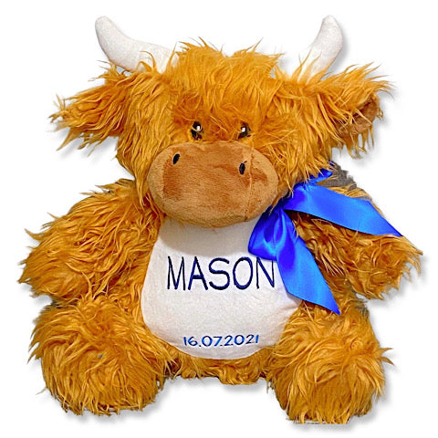 Shaggy brown highland cow plushie teddy with white horns personalised with a name and date of birth embroidered in shades of blue. A dark blue ribbon has been tied in a bow around the neck.